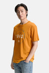 T-shirt ocre à manches courtes de Tom Tailor, avec imprimé graphique centré, porté avec un jean décontracté.

