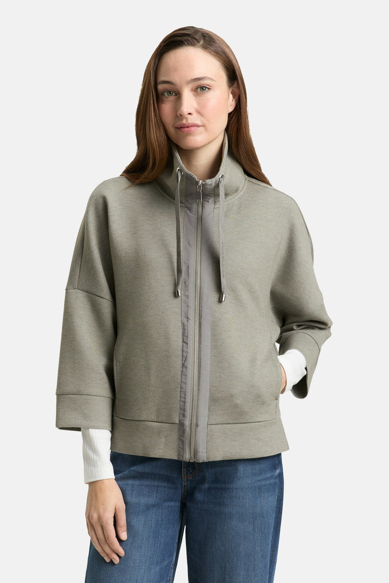 Cardigan kaki de Tom Tailor, avec fermeture éclair et manches trois quarts, porté sur un pull blanc et un jean.
