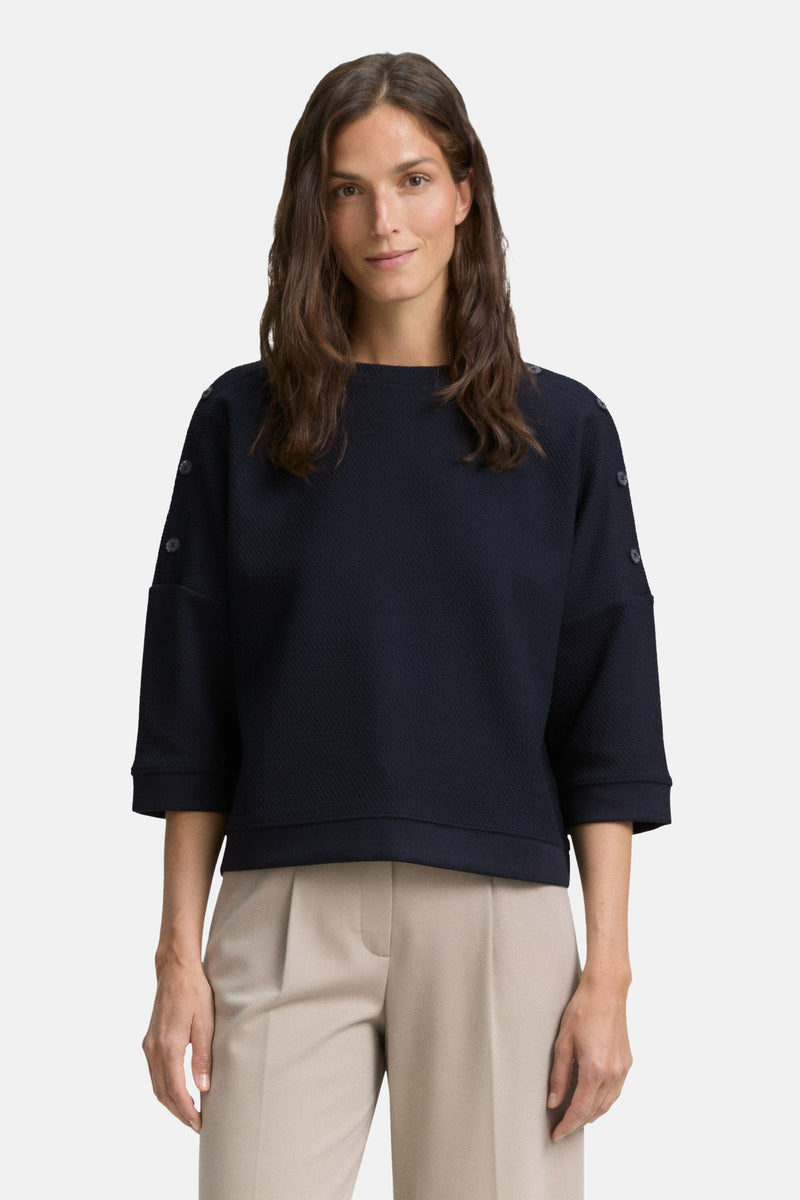 Blauwe sweater van Tom Tailor, driekwartmouwen met decoratieve knopen, gedragen met een effen beige broek.