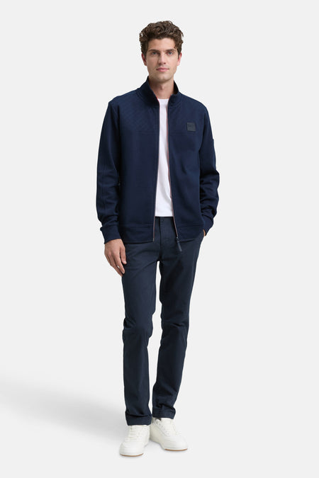Blauwe Tom Tailor jas met ritssluiting, ruitpatroon aan de bovenkant en gedragen over een wit T-shirt en een marineblauwe chino.