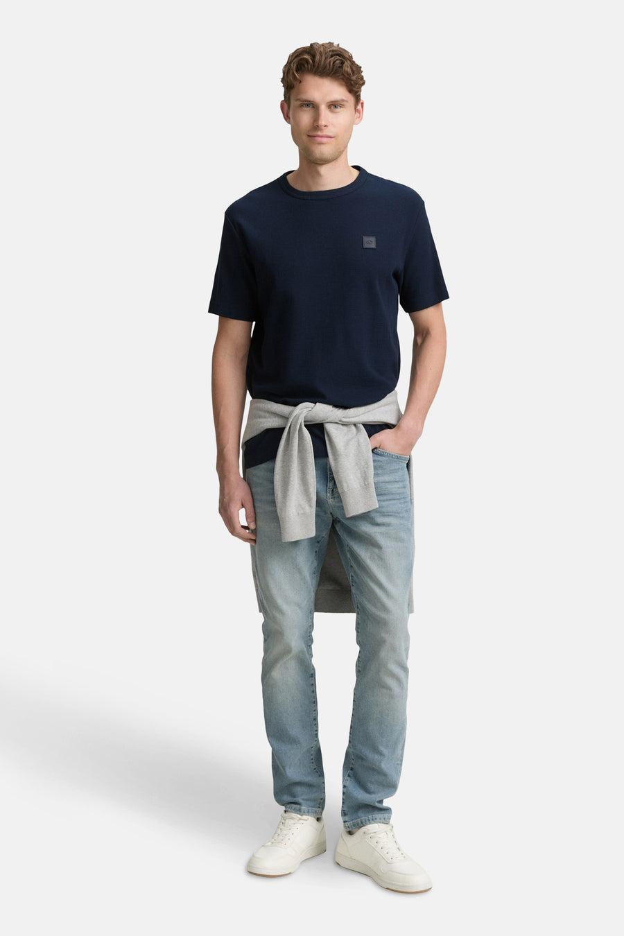 Blauw T-shirt met korte mouwen van Tom Tailor, gedragen met een grijze trui die om de schouders is geknoopt en een lichtblauwe jeans.
