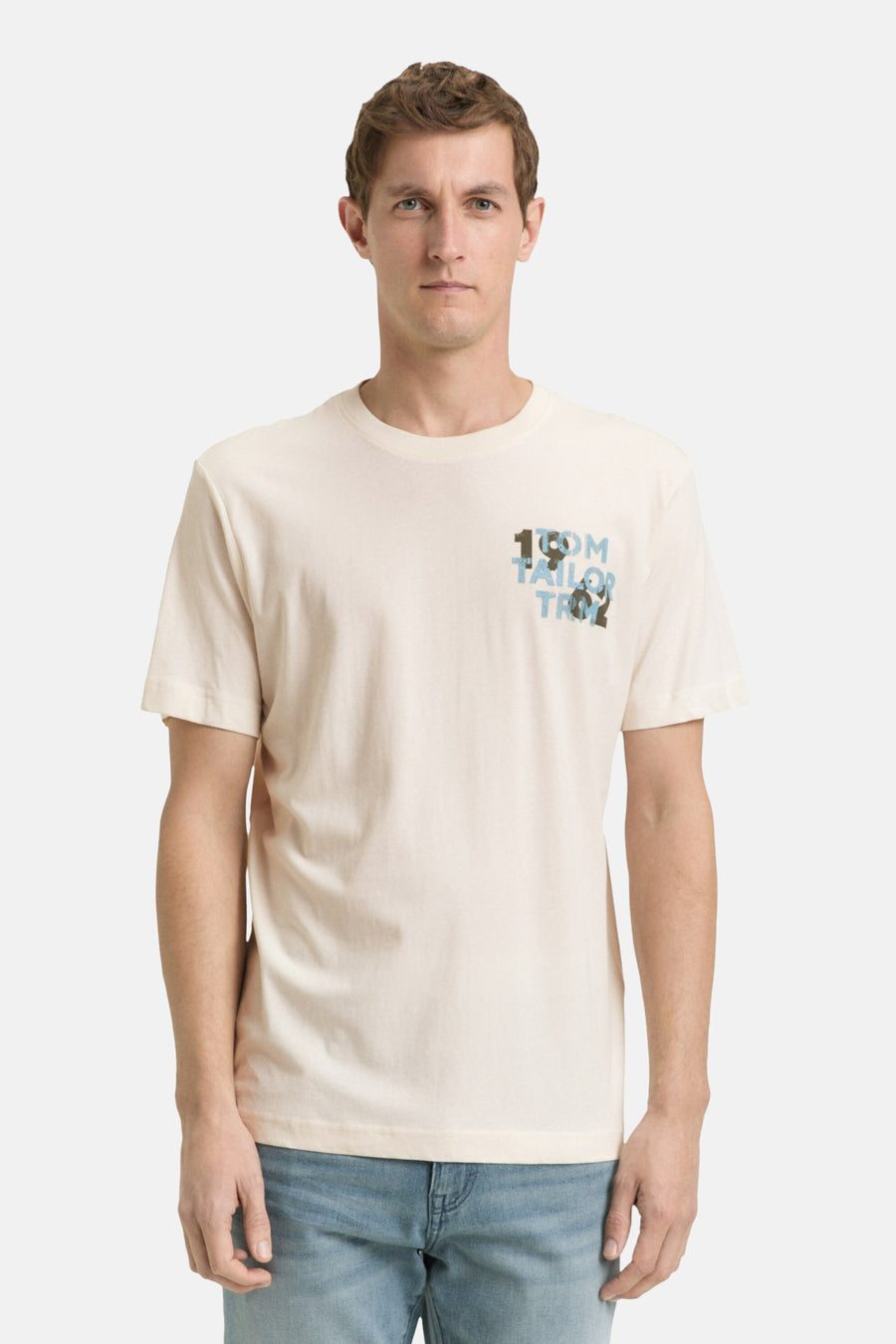 Ecru T-shirt met korte mouwen van Tom Tailor, met een decoratief logo op de borst, gedragen met een blauwe jeans en witte sneakers.