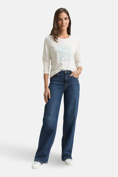T-shirt à manches longues blanc de Tom Tailor, avec un imprimé graphique sur le devant, porté avec un jean bleu et des chaussures blanches.