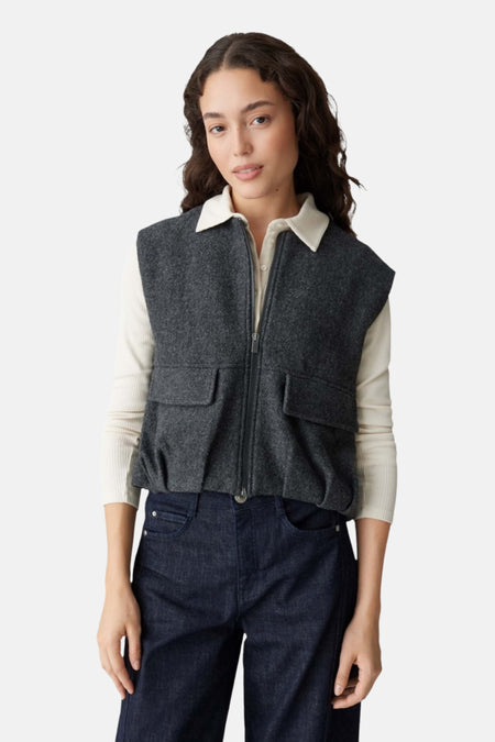 Veste grise par OPUS avec fermeture éclair, poches et détails plissés portée sur un haut blanc et un pantalon en denim foncé.