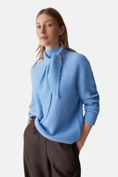 Pull bleu d'OPUS avec un col montant à nouer et manches longues, porté avec un pantalon marron.