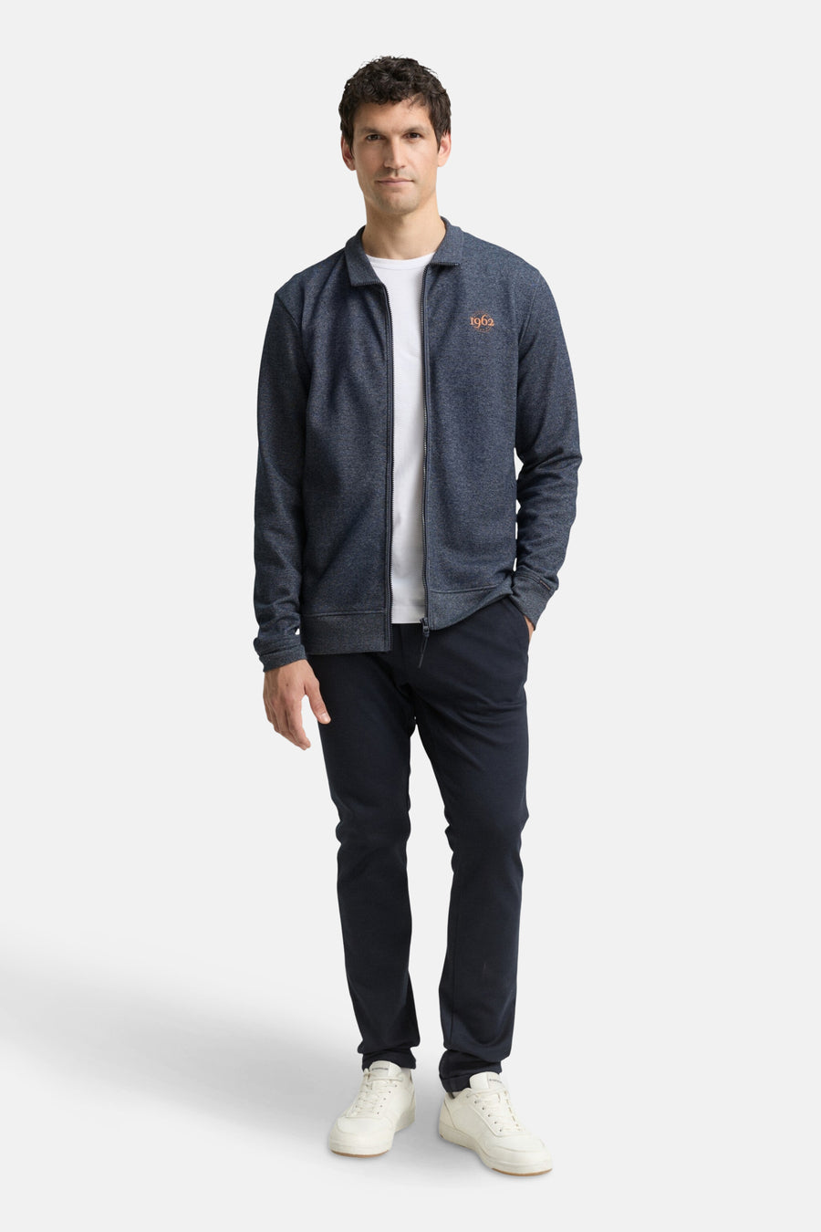Gilet bleu de Tom Tailor, avec fermeture éclair et inscription « 1962 », porté sur un t-shirt blanc et un pantalon bleu marine.
