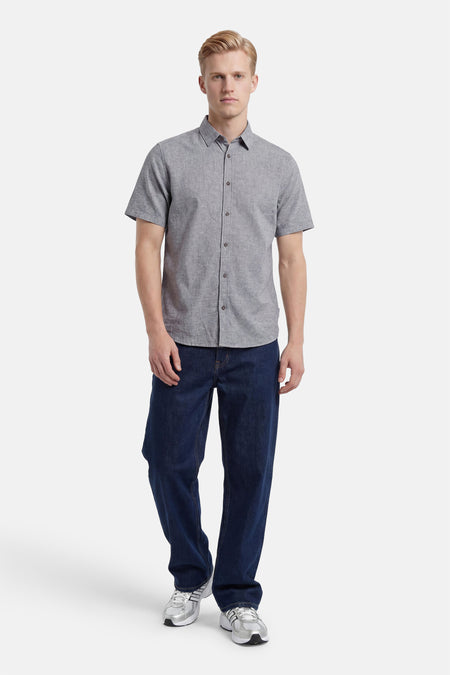 Chemise à manches courtes grise de Tom Tailor, texture chinée, avec boutons foncés, portée par un homme avec un jean brut et des baskets bicolores.