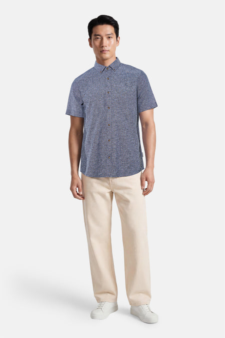 Chemise bleue à manches courtes de Tom Tailor, en tissu texturé chiné, portée par un homme avec un pantalon beige et des baskets blanches.
