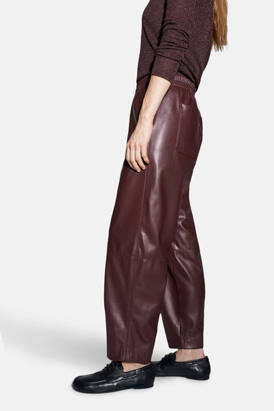 Pantalon bordeaux en similicuir par OPUS, avec une taille élastique et porté avec un pull brillant et des chaussures bateau noires.