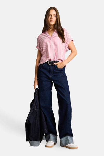 Chemisier à manches courtes multicolore de Tom Tailor, rose à rayures blanches, rentré dans un jean large brut, tenu d'une main.