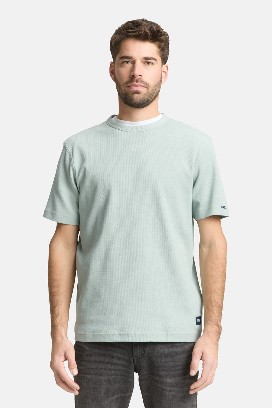 T-shirt à manches courtes vert de Tom Tailor, avec texture côtelée et ourlet droit, porté avec un t-shirt blanc à col rond et un jean gris.