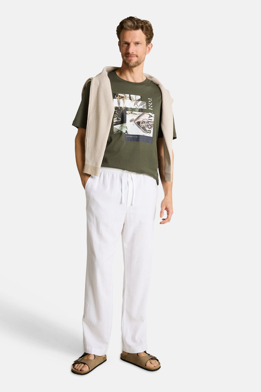 T-shirt vert à manches courtes de Tom Tailor avec un imprimé graphique, porté par un homme avec un pull beige sur les épaules, un pantalon blanc et des sandales.