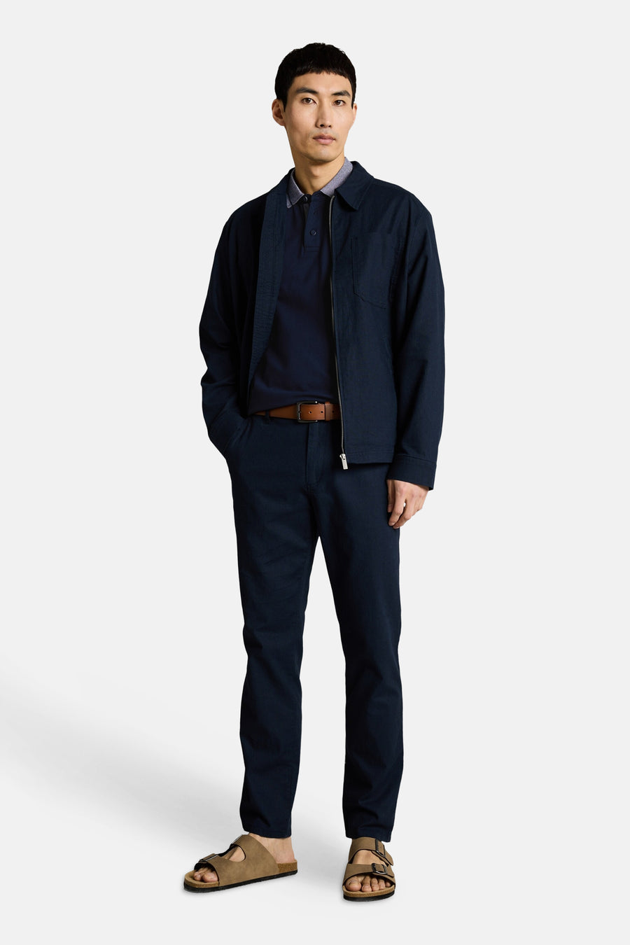 Polo bleu marine de Tom Tailor à col zippé, porté avec veste, pantalon assorti, ceinture marron et sandales.