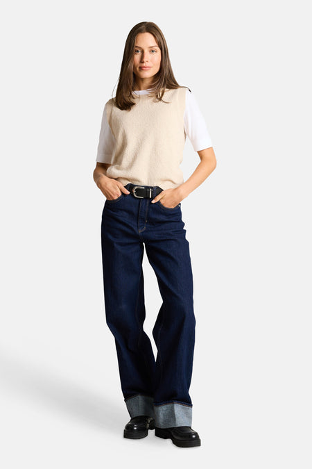 Top sans manches beige de Tom Tailor, porté sur un t-shirt blanc avec un jean taille haute bleu foncé et une ceinture noire.