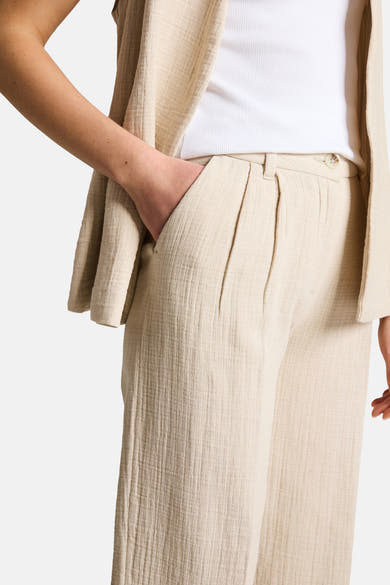Pantalon beige de Tom Tailor en tissu froissé, avec pinces et bouton apparent, main dans la poche.
