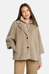 Taupe faux fur poncho with wide collar and black buttons, worn over ribbed sweater and beige pants, front view