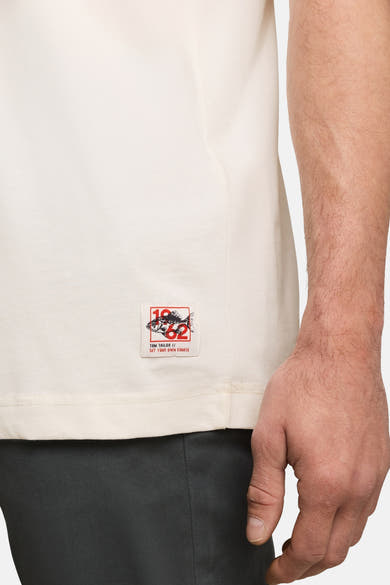 White short-sleeve T-shirt by Tom Tailor with red and black logo patch on lower left side, worn with dark trousers, side view close-up.