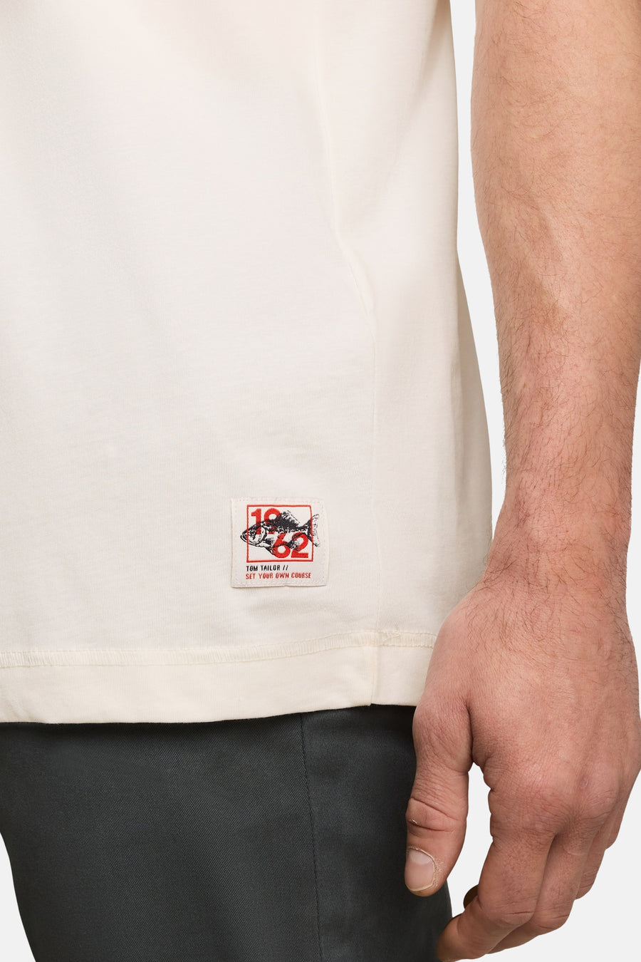 White short-sleeve T-shirt by Tom Tailor with red and black logo patch on lower left side, worn with dark trousers, side view close-up.