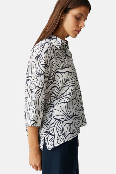 Chemisier 3/4 blanc à motifs feuillage foncé de OPUS, style chemise boutonnée, vu de côté avec pantalon foncé.