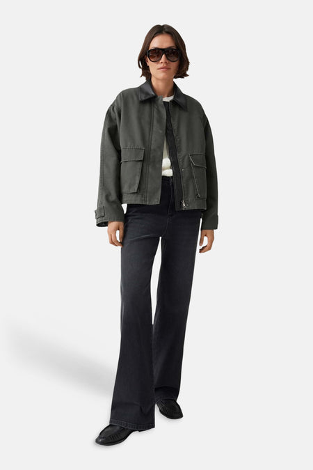 Olive green parka with front pockets and button closure, worn by model in full-length lifestyle view with dark jeans and black loafers.