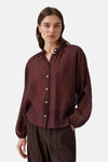 Bordeaux long-sleeve blouse with gathered neckline and white buttons, featuring a relaxed fit and textured fabric, front view on model wearing dark pleated trousers