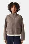 Taupe bomber jacket with front zip and two patch pockets, worn over white ribbed top and dark jeans, front view.