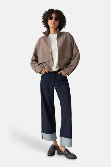 Taupe bomber jacket with front zip and flap pockets, worn over white ribbed top, paired with dark blue wide-leg jeans and brown loafers, full-length lifestyle view