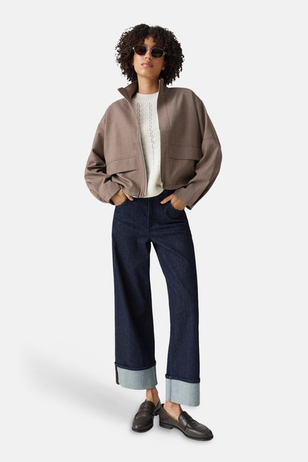 Taupe bomber jacket with front zip and flap pockets, worn over white ribbed top, paired with dark blue wide-leg jeans and brown loafers, full-length lifestyle view