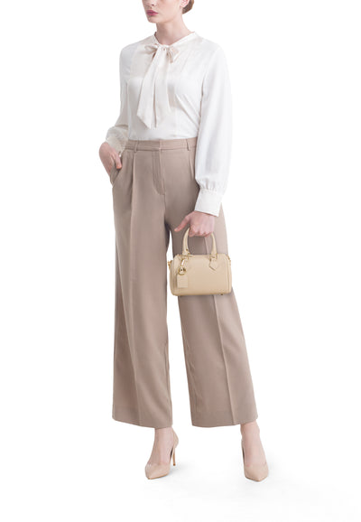 Ecru textuurige handtas van Access® met bovenhandvat en bedel, gedragen door een vrouw in een witte blouse en een beige broek.