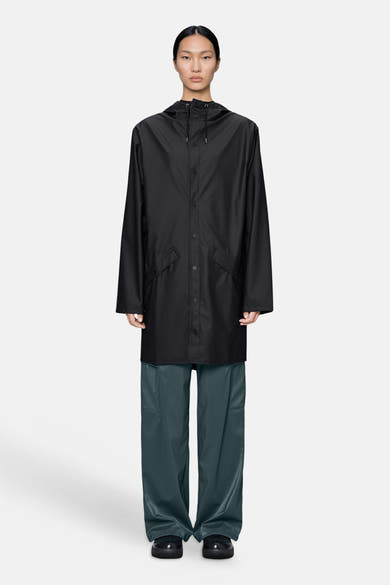 Black long coat with hood and front buttons, worn by model in full-length studio view, paired with dark green trousers and black shoes.