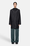 Black long coat with hood and front buttons, worn by model in full-length studio view, paired with dark green trousers and black shoes.