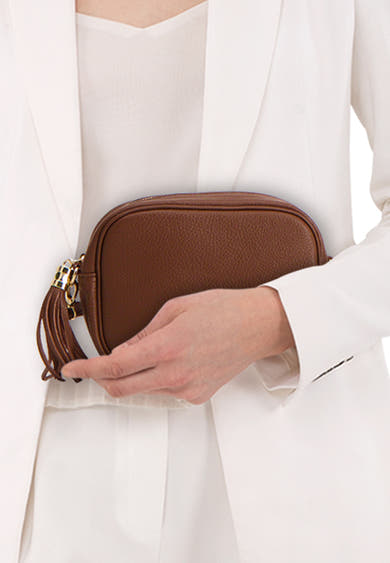 Sac à bandoulière brun par Access®, avec une chaîne dorée et une frange décorative, porté avec un tailleur blanc.