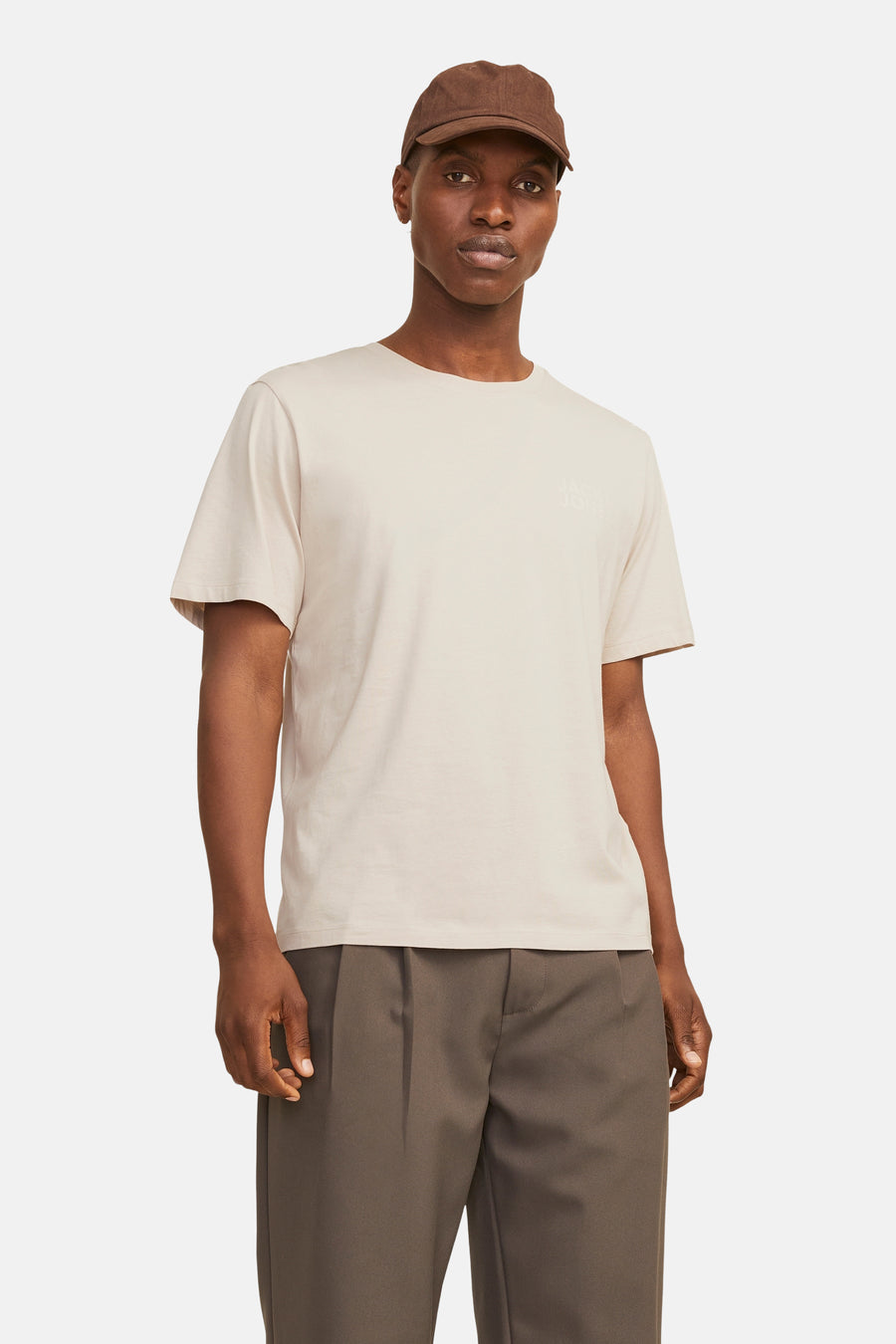 T-shirt beige à manches courtes de JACK & JONES, porté avec un pantalon marron plissé et une casquette.
