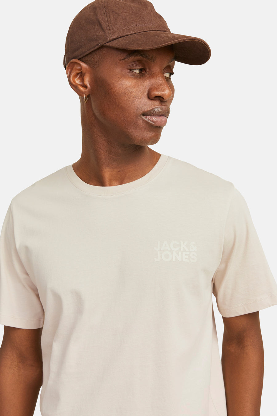 T-shirt beige à manches courtes de JACK & JONES, porté avec une casquette marron et une boucle d'oreille dorée.