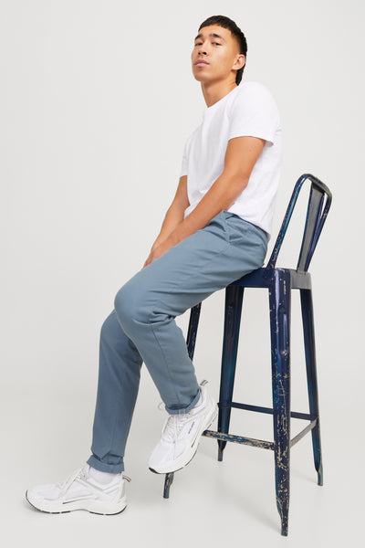 Blauwe chino van JACK & JONES, gedragen met een wit T-shirt en witte sneakers op een blauwe kruk.