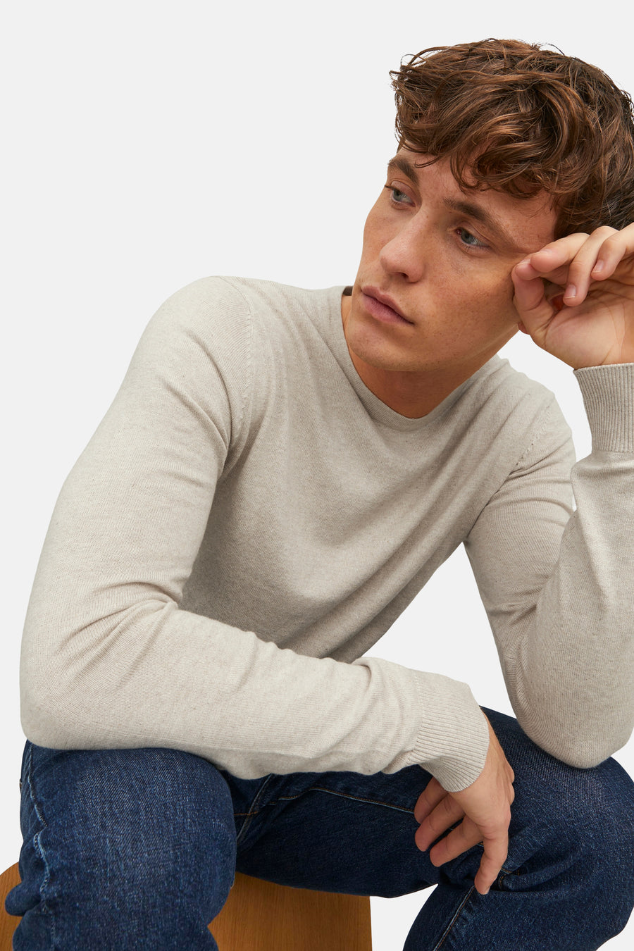 Pull beige de PREMIUM BLUE by JACK & JONES, porté avec un jean bleu foncé, assis sur une chaise.