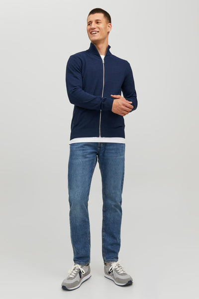 Navy blue knit gilet with full zipper and ribbed cuffs, worn over white tee with jeans and gray sneakers, front view.