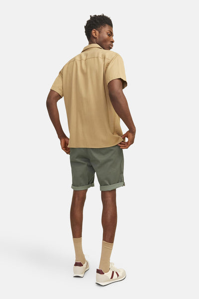 Short olive de JACK & JONES JEANS INTELLIGENCE, avec revers, porté avec une chemise beige et des baskets rétro.