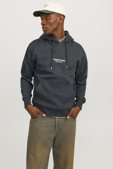Dark green hooded sweatshirt by Originals Studio with white chest logo, front kangaroo pocket, and drawstrings, worn with olive jeans and a cream cap.