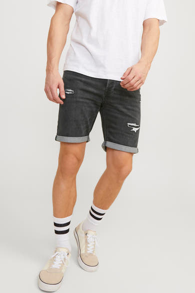 Korte zwarte spijkerbroek van JACK & JONES JEANS INTELLIGENCE, met gescheurde details en opgerolde zomen, gedragen met een wit T-shirt en gestreepte sokken.
