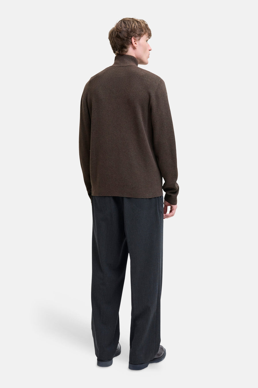 Brown turtleneck sweater by [brand omitted] shown from back, ribbed texture, long sleeves, paired with dark trousers and black shoes.