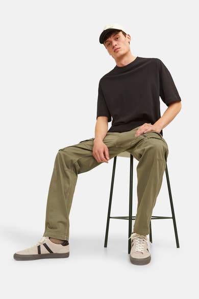 Kaki cargobroek van JACK & JONES, gedragen met een zwart T-shirt en beige sneakers, man zittend op een stoel.