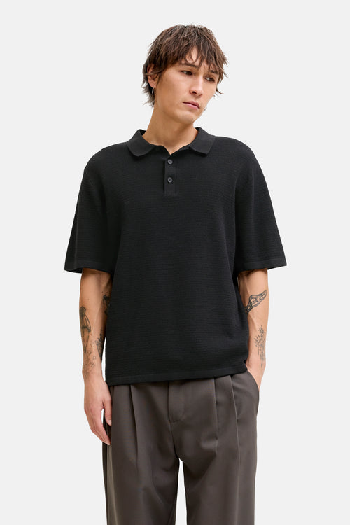 Zwarte, getextureerde polo van JACK & JONES, gedragen met een wijde grijze broek, korte mouwen en een relaxte pasvorm.