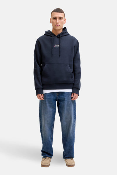 Sweat à capuche bleu marine par JACK & JONES, porté sur un t-shirt blanc et un jean décontracté, avec des chaussures couleur sable.
