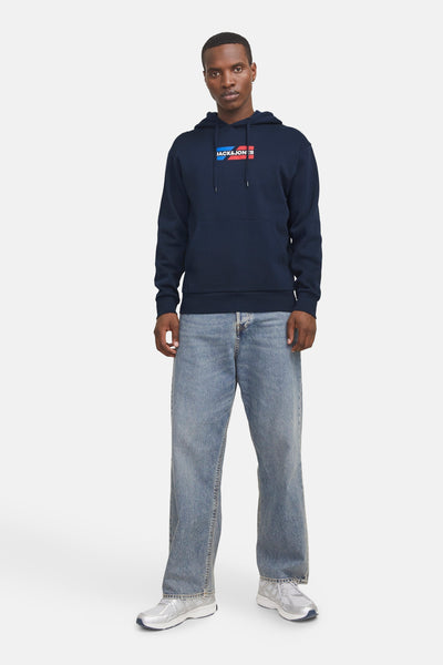Marineblauwe hoodie van JACK & JONES, met een rechthoekig logo op de borst, gedragen met een wijde jeans en zilveren sneakers.