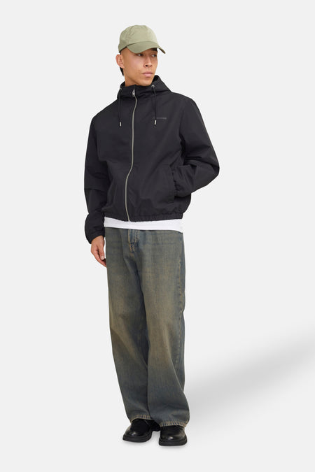 Zwarte regenjas van ORIGINALS BY JACK & JONES, met capuchon en ritssluiting, gedragen met een wit T-shirt en een wijde jeans.