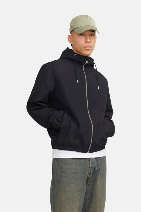 Zwarte regenjas van ORIGINALS BY JACK & JONES, met capuchon en ritssluiting, gedragen over een wit T-shirt en een spijkerbroek.