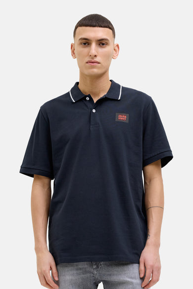 Donkerblauwe polo met korte mouwen van JACK & JONES, met vierkant logo op de borst, gedragen met een spijkerbroekshort.