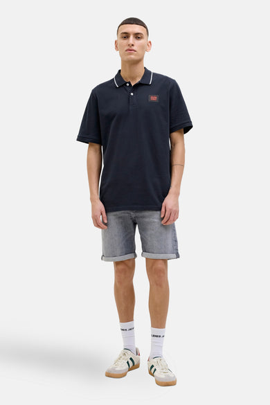 Polo à manches courtes bleu foncé de JACK & JONES, avec logo carré sur la poitrine, porté avec un short en jean.