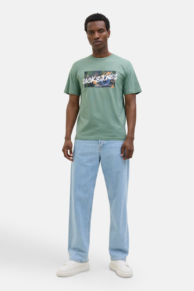 Kaki T-shirt met korte mouwen van JACK & JONES, met grafische print op de voorkant, gedragen met lichtblauwe jeans en witte sneakers.